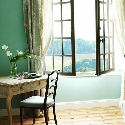 Blue Bedroom with open window, blue walls, blue and white floral curtains, desk with flowers and magazine.