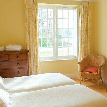 Bedroom with twin beds, yellow walls, yellow floral curtains, wooden drawers and table.