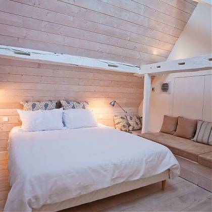 Bedroom with double bed, white bedlinen, wooden panels on walls and floors, seating area on right side.