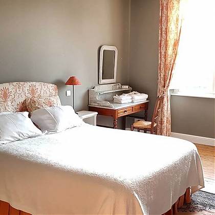 Bedroom with double bed, white bedspread, peach and white floral curtains and headboard, bedside table with towels.