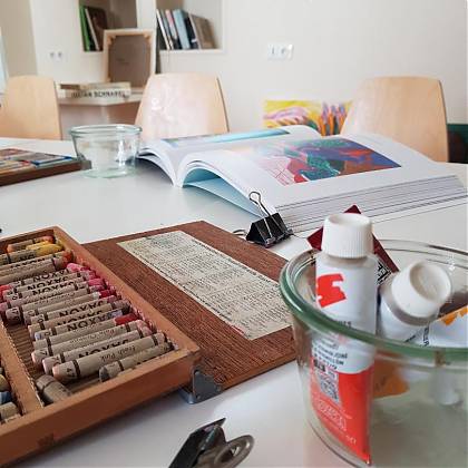 Box of pastel crayons, tubes of paint, art book on table.
