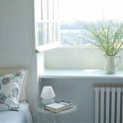 Bedroom with bed and pillow, side table with book and lamp, open window with green flowers in white jug.
