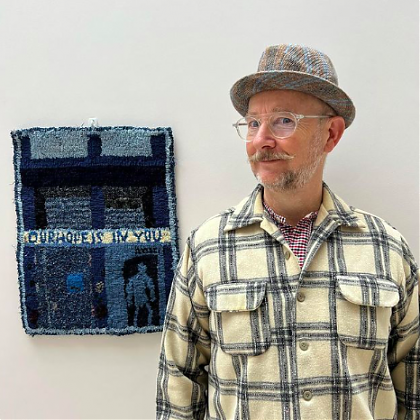 Man in hat with checked shirt, in front of a hooked rug on wall.