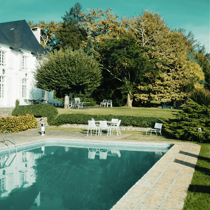 Swimming pool with terrace, tables and chairs. White manor house with black roof.